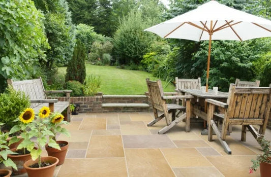 A stone patio with paving tiles, a wooden table and chairs, an open white umbrella, potted sunflowers, and a lush green garden with trees and grass in the background.