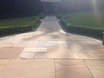 A stone pathway with large buff sandstone paving tiles leads through a manicured lawn bordered by hedges toward a wooden gazebo in the background. Sunlight creates soft shadows on the path.