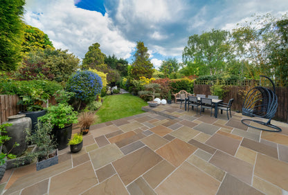 Spacious backyard with a stone patio featuring Buff Sandstone Paving, an outdoor dining table, hanging chair, potted plants, and a lush green lawn bordered by trees, shrubs, and colorful flowers under a partly cloudy sky.