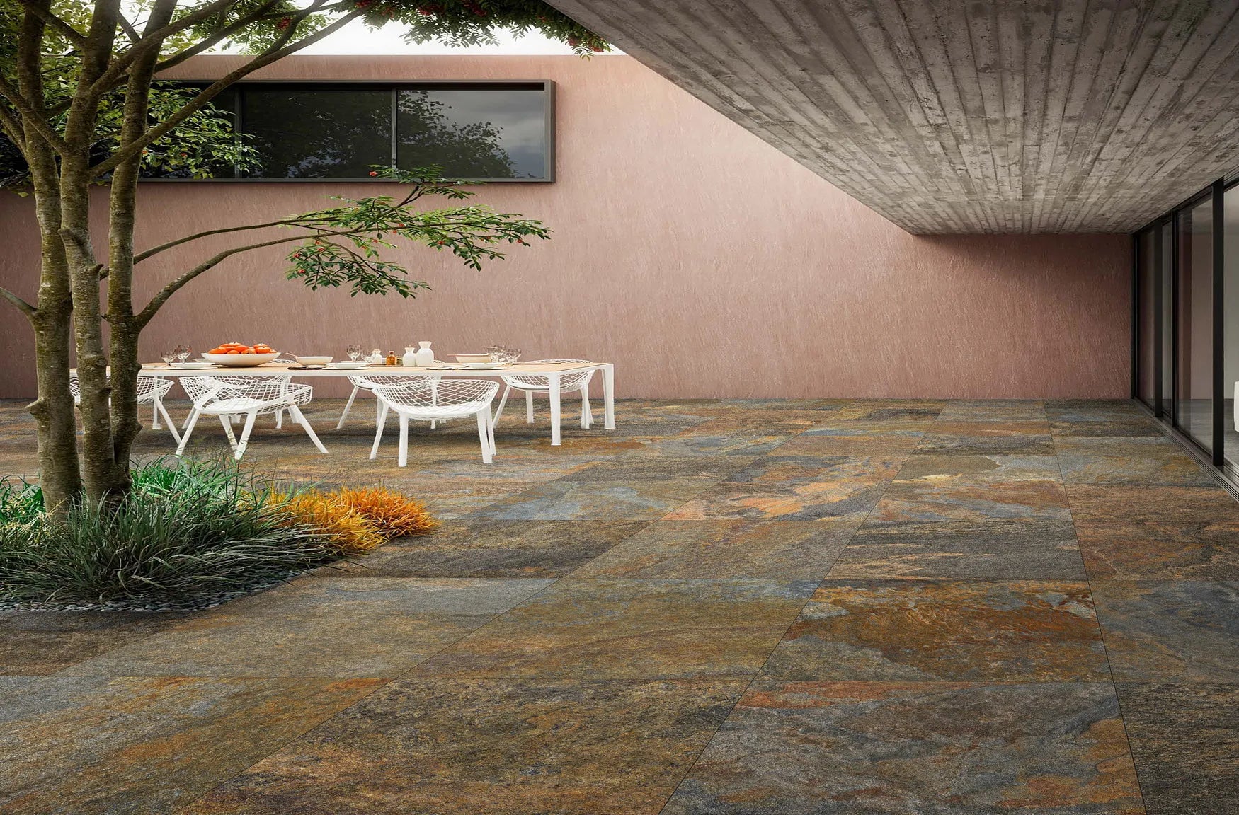 A modern outdoor patio features slip-resistant paving tiles, a white dining table and chairs, a small garden area with plants and a tree, plus a building with large windows and an overhanging concrete roof.