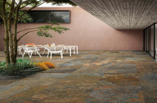 A modern outdoor patio features slip-resistant paving tiles, a white dining table and chairs, a small garden area with plants and a tree, plus a building with large windows and an overhanging concrete roof.