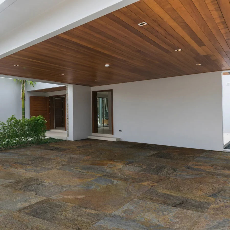 A modern open garage with a wooden ceiling, white walls, large windows, and a floor made of large, textured stone tiles. Green shrubs are visible along one side near the entrance.
