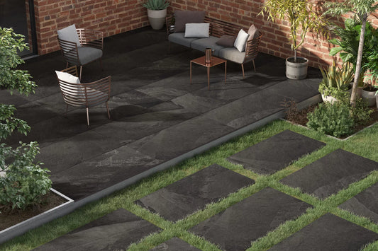 Modern outdoor patio with large Falkland Porcelain Paving Slabs, surrounded by red brick walls, potted plants, contemporary metal chairs, a small table, and a lower area with porcelain paving tiles set in green grass.