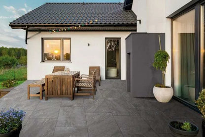 A modern outdoor patio with grey porcelain paving, wooden dining table and chairs, potted plants, and string lights; the house has white walls and large windows, with a sunset reflecting in the window.