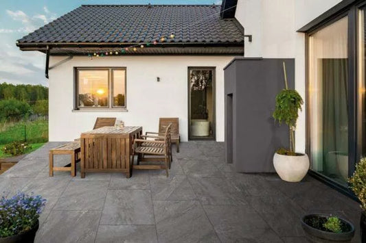 A modern outdoor patio with grey porcelain paving, wooden dining table and chairs, potted plants, and string lights; the house has white walls and large windows, with a sunset reflecting in the window.