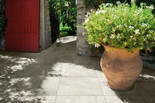 A large terracotta pot with green and white flowering plants sits on porcelain paving tiles, next to a stone wall and a partially open red wooden door, with sunlight and leafy shadows on the ground.