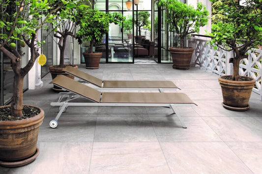 Two empty brown lounge chairs sit side by side on a tiled patio paved with Fraser Grigio Porcelain Paving Slabs, surrounded by large potted trees, plants, and glass doors leading to more seating in the background.