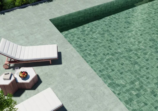 Two white cushioned lounge chairs and a small side table with fruit and a drink are set beside a clear green-tiled swimming pool, surrounded by elegant French Gris porcelain tile on a sunny day.