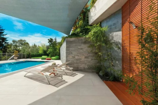 Modern patio with two white lounge chairs beside a swimming pool, surrounded by greenery and trees. Porcelain paving adds a sleek touch, while a stone wall and wooden slat screen provide privacy under bright sunlight and shifting shadows.