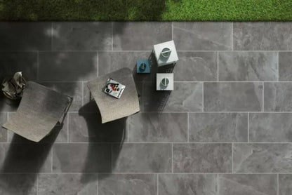 Two modern gray chairs and a small table with decorative items sit on durable pavers next to a patch of green grass. Shadows from objects and foliage create striking patterns across the gray stone-tiled patio.