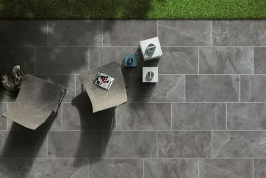 Two modern gray chairs and a small table with decorative items sit on durable pavers next to a patch of green grass. Shadows from objects and foliage create striking patterns across the gray stone-tiled patio.