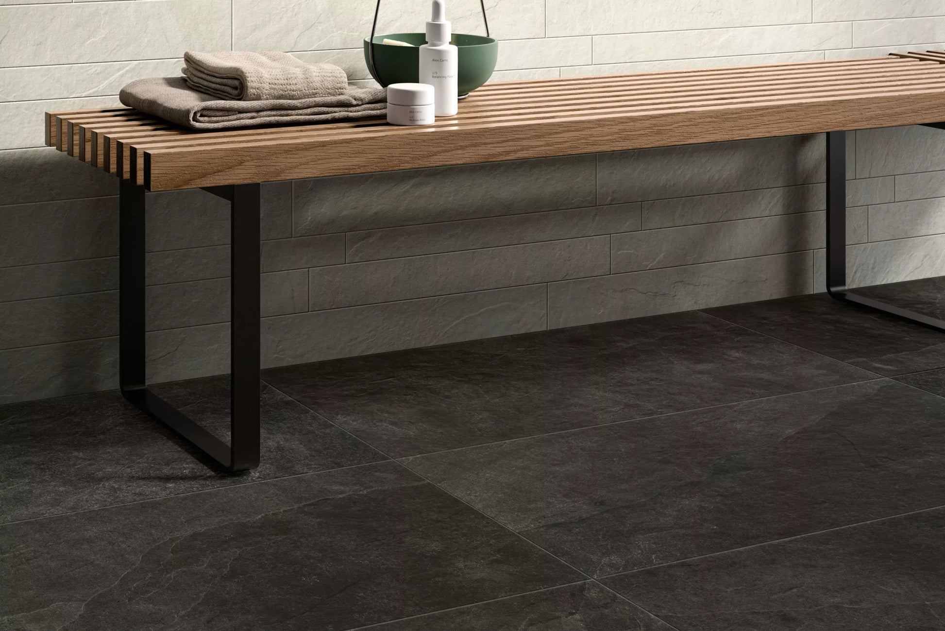 A wooden bench with metal legs sits against a light stone-textured wall; folded towels and toiletries are placed on top. The floor features large, dark porcelain paving slabs for a modern, durable touch.