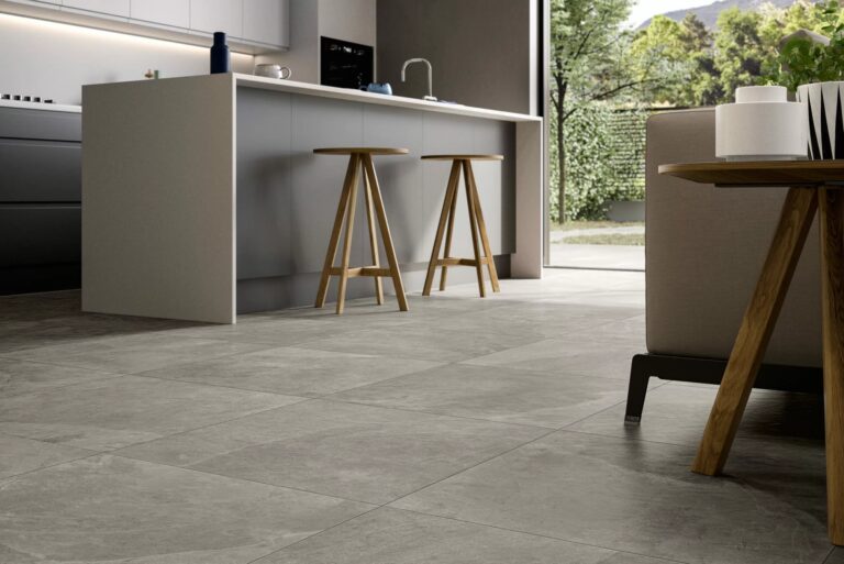 Modern kitchen with light gray cabinets, a central island with two wooden stools, large gray stone tile flooring that echoes the Fuego River Porcelain Paving seen outside, minimal decor, and a view of greenery through a glass door.