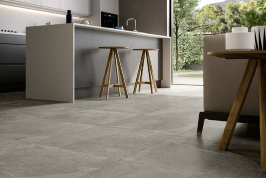 Modern kitchen with light gray cabinets, a central island with two wooden stools, large gray stone tile flooring that echoes the Fuego River Porcelain Paving seen outside, minimal decor, and a view of greenery through a glass door.