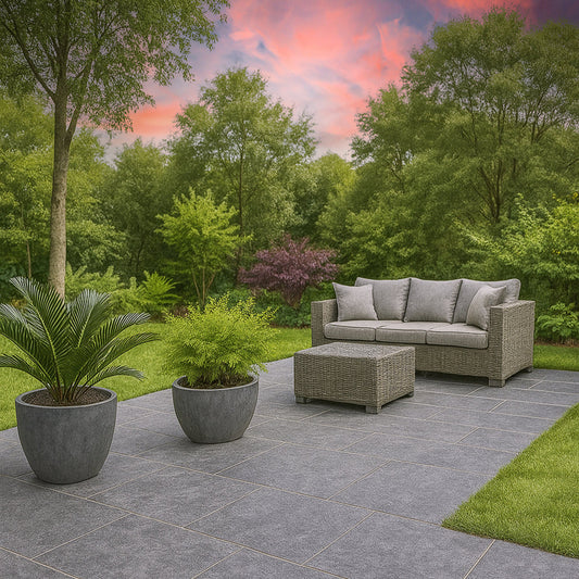 A modern patio with slip-resistant, gray porcelain tiles features a wicker sofa with gray cushions, a matching coffee table, and two large potted plants. The area is surrounded by lush greenery and trees under a colorful sunset sky.