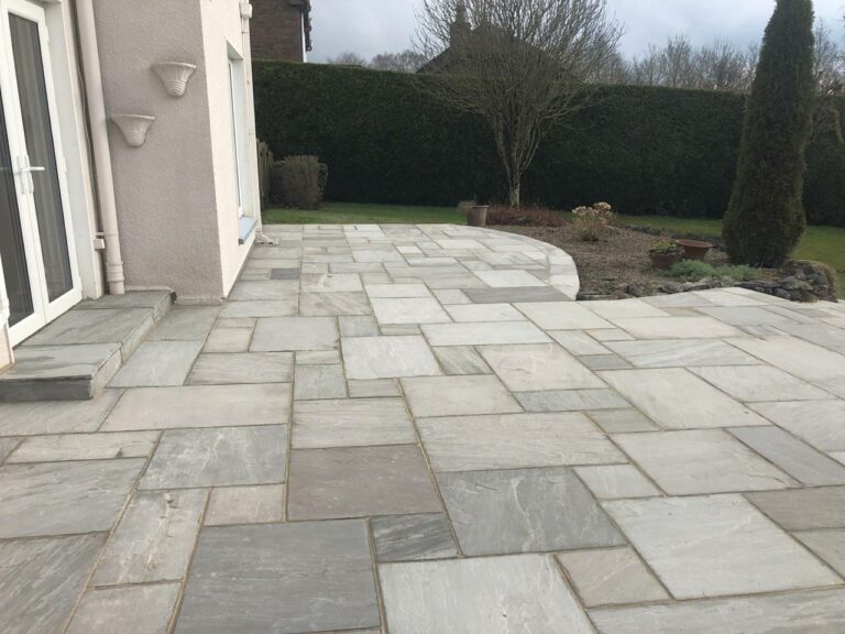 A spacious outdoor patio with light gray, rectangular stone tiles, adjacent to a house. The patio curves toward a garden area with trimmed grass, a few trees, and shrubs along the edge.