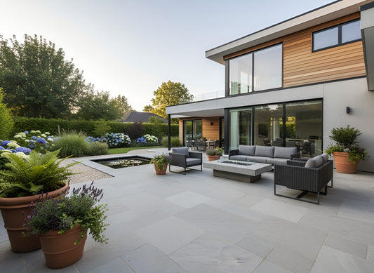 Modern backyard patio with Kandla Grey Sandstone Paving tiles, outdoor seating around a fire table, potted plants, a small pond, and a contemporary house with large windows in the background.