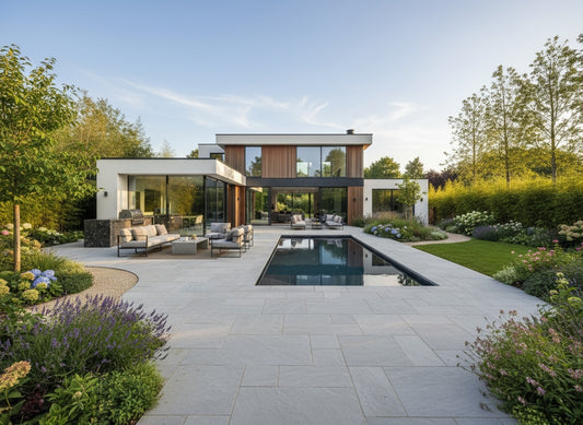 Modern house with large glass windows, wooden accents, and flat roof, surrounded by lush gardens. A rectangular pool with lounge seating lines the stone patio, featuring elegant Kandla Grey Sandstone Paving, on a clear, sunny day.