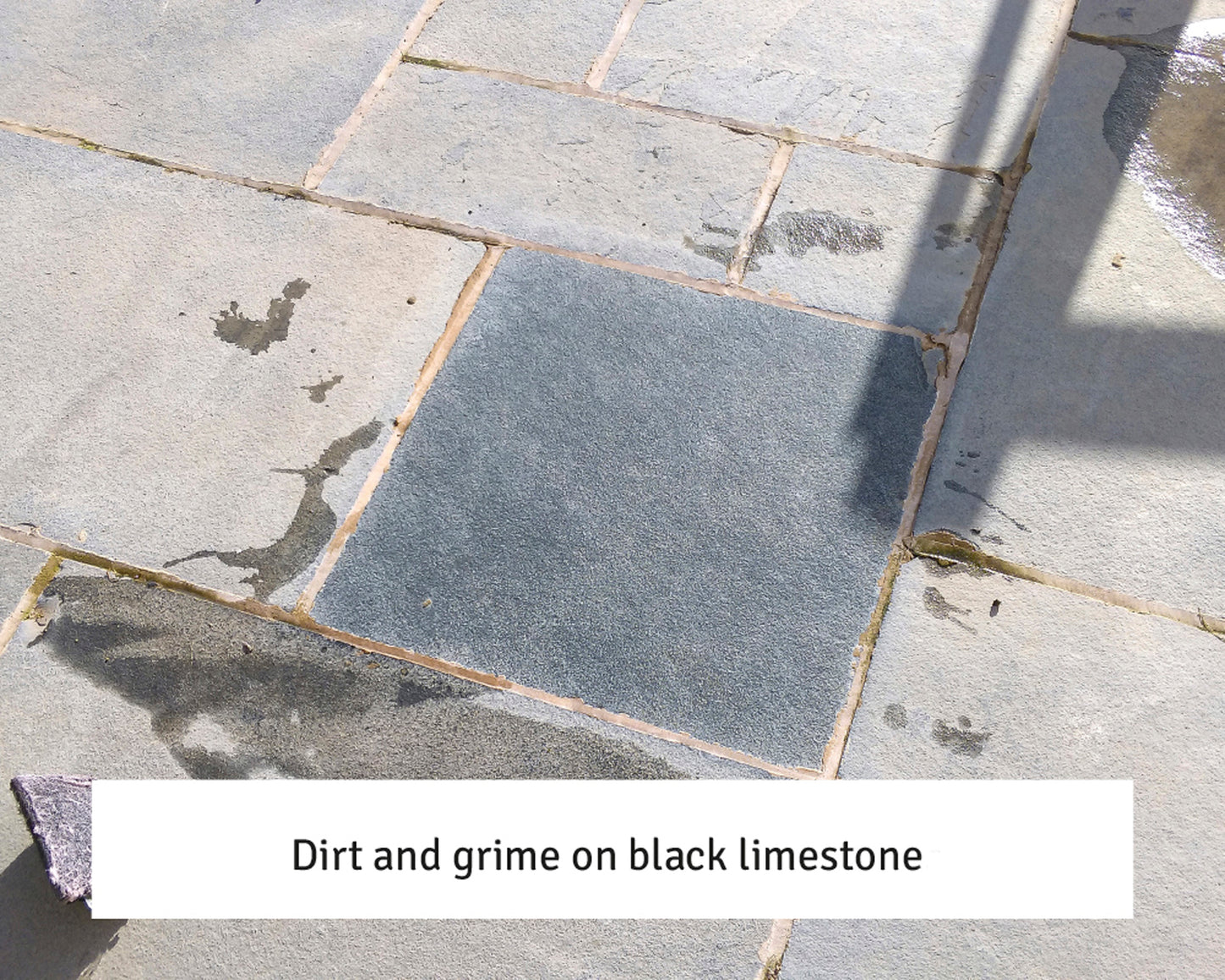 A patio made of light-colored stone slabs with a central dark black limestone tile showing visible dirt and grime, surrounded by shadows and water stains. Text at the bottom reads, "Dirt and grime on black limestone – try a stone cleaner.