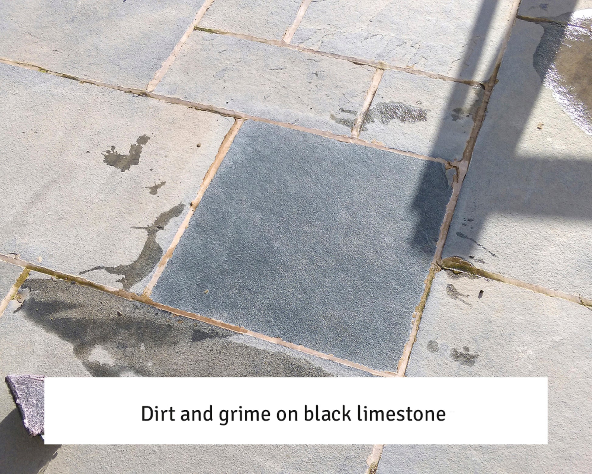 A patio made of light-colored stone slabs with a central dark black limestone tile showing visible dirt and grime, surrounded by shadows and water stains. Text at the bottom reads, "Dirt and grime on black limestone – try a stone cleaner.