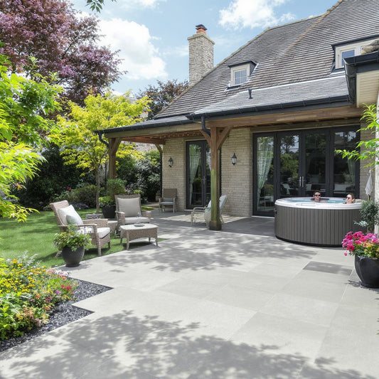 Spacious patio with light gray Palmer Grey Porcelain Paving Slabs, outdoor furniture, potted plants, and a round hot tub. Two people relax in the tub near a modern house with a covered porch, surrounded by lush greenery and flowers.