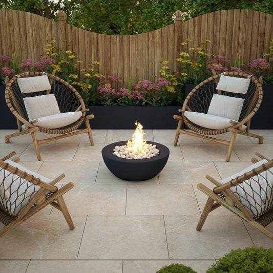 A modern patio with four wooden lounge chairs surrounds a black fire pit with flames, set on slip-resistant paving tiles. A wooden fence and blooming flowers complete the stylish outdoor setting.