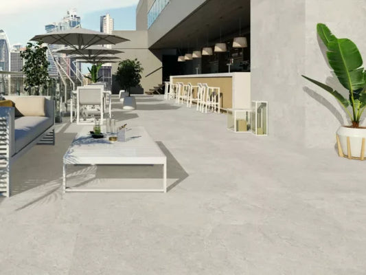 A modern rooftop terrace with white outdoor furniture, potted plants, an umbrella, and a bar area with stools features slip-resistant paving tiles and overlooks a city skyline under a clear sky.