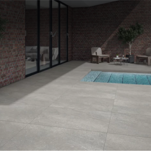 A modern patio with light gray stone tiles, outdoor seating, potted plants, and a small rectangular pool next to large glass doors and brick walls.