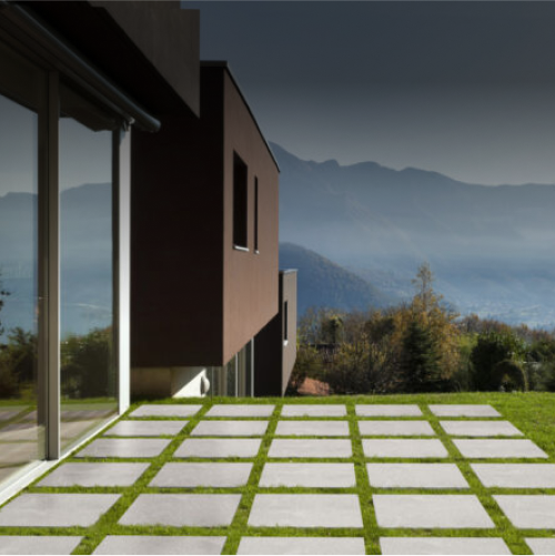 Modern house with large glass windows and a geometric patio featuring square stone tiles in a grid pattern with grass between them, overlooking a scenic mountain landscape under a cloudy sky.