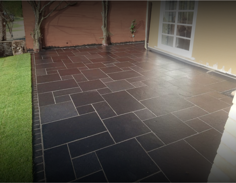 A clean, wet, dark stone patio with rectangular tiles, bordered by grass on one side and a yellow house with glass doors on the other. A few bare trees and plants are near the walls.