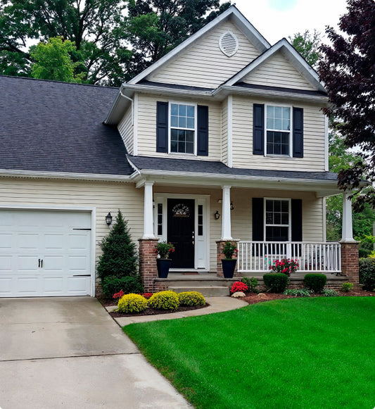 A two-story beige house with black shutters, a front porch, potted plants, and manicured landscaping. A concrete driveway leads to a white garage, and the yard features green grass and neatly trimmed bushes.