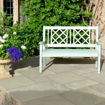 A light green wooden bench sits on a stone patio of mint sandstone paving, surrounded by lush green plants and flowers, including a pot of vibrant purple blooms on the left.
