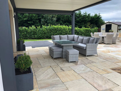 A covered outdoor patio with a gray wicker sectional sofa, glass-top table, and matching stools rests on Mint Sandstone Paving tiles. In the background, a dining set and greenery border the stylish space.