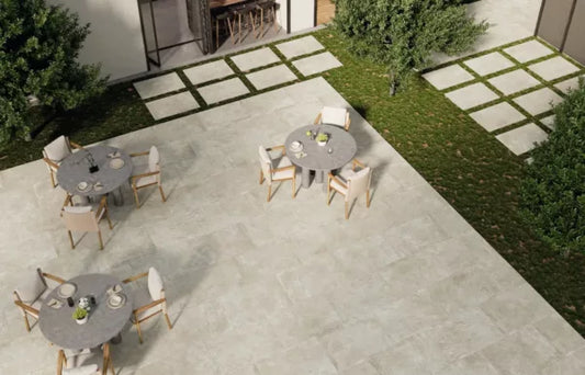 A modern outdoor patio with Palmer Bone Porcelain Paving Slabs, round tables with chairs, and green grass sections featuring square stepping stones. Trees and shrubs border the area, creating a serene setting.