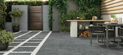 Modern outdoor patio with stylish Palmer Graphite Porcelain Paving Slabs, a dining table with woven chairs, potted plants, a small outdoor kitchen with wood storage, and lush greenery along the fence and walls.