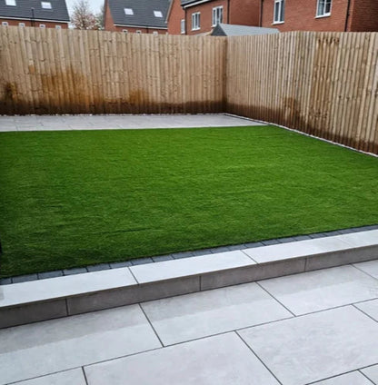 A neatly landscaped backyard with a rectangular patch of green grass bordered by Palmer Grey Porcelain Paving Slabs and a wooden fence. Brick houses are visible in the background, adding charm to this inviting outdoor space.
