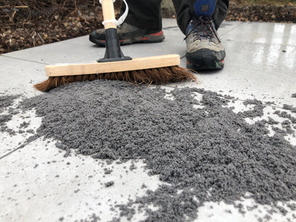 A person wearing outdoor shoes sweeps gray sand or fine gravel, possibly as a brush-in jointing solution, across large stone tiles with a broom on a patio or sidewalk.