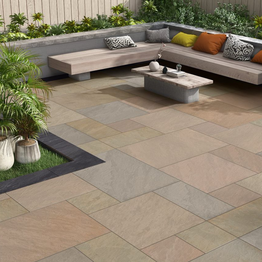 Modern outdoor patio featuring Raj Green Porcelain Paving Slabs in beige and light brown, a corner wooden bench with colorful cushions, a low rectangular coffee table, lush potted plants, and a raised planter bed along a wooden fence.