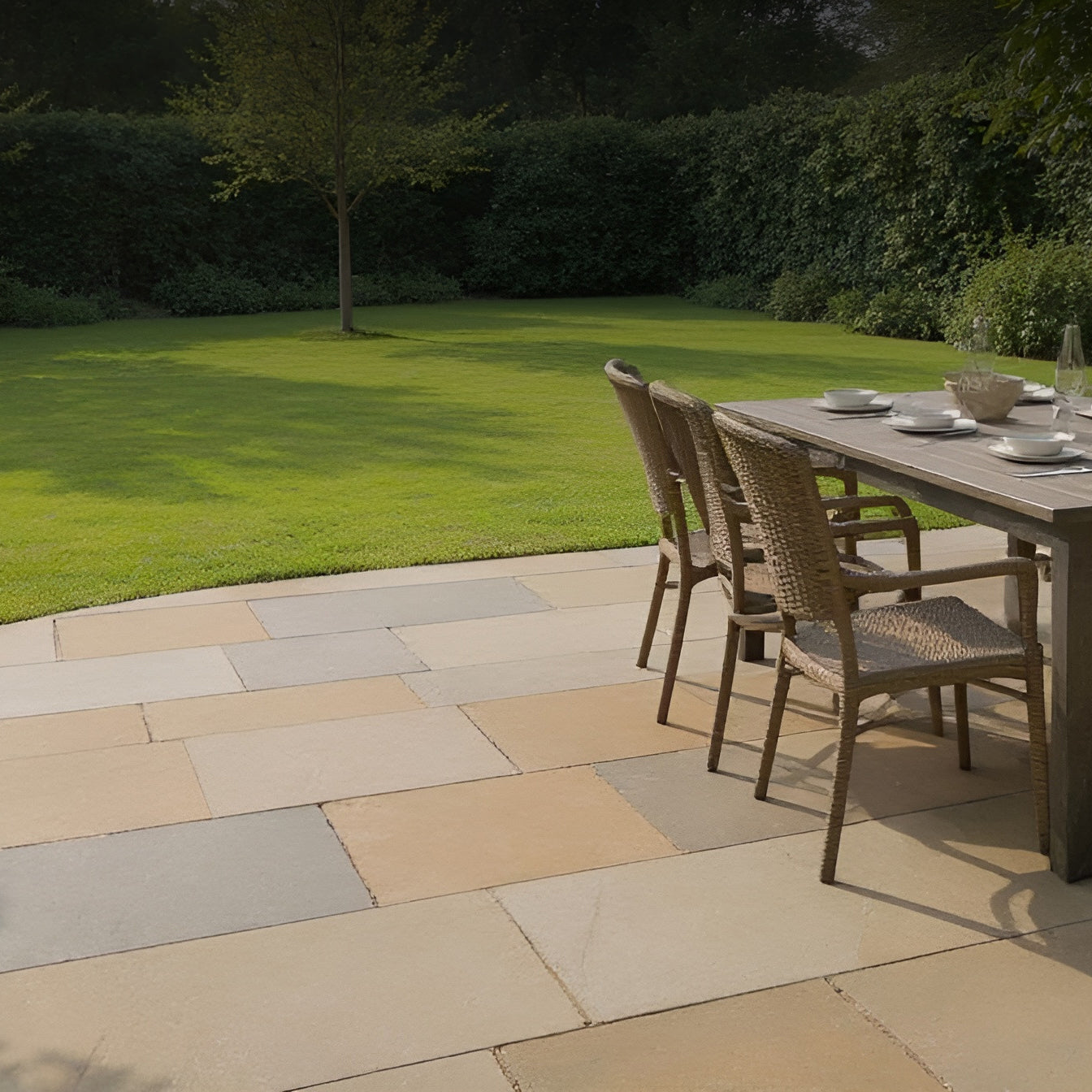 A patio with wicker chairs and a dining table set for four, overlooking a neatly mowed lawn bordered by dense green bushes and a single tree in the background.