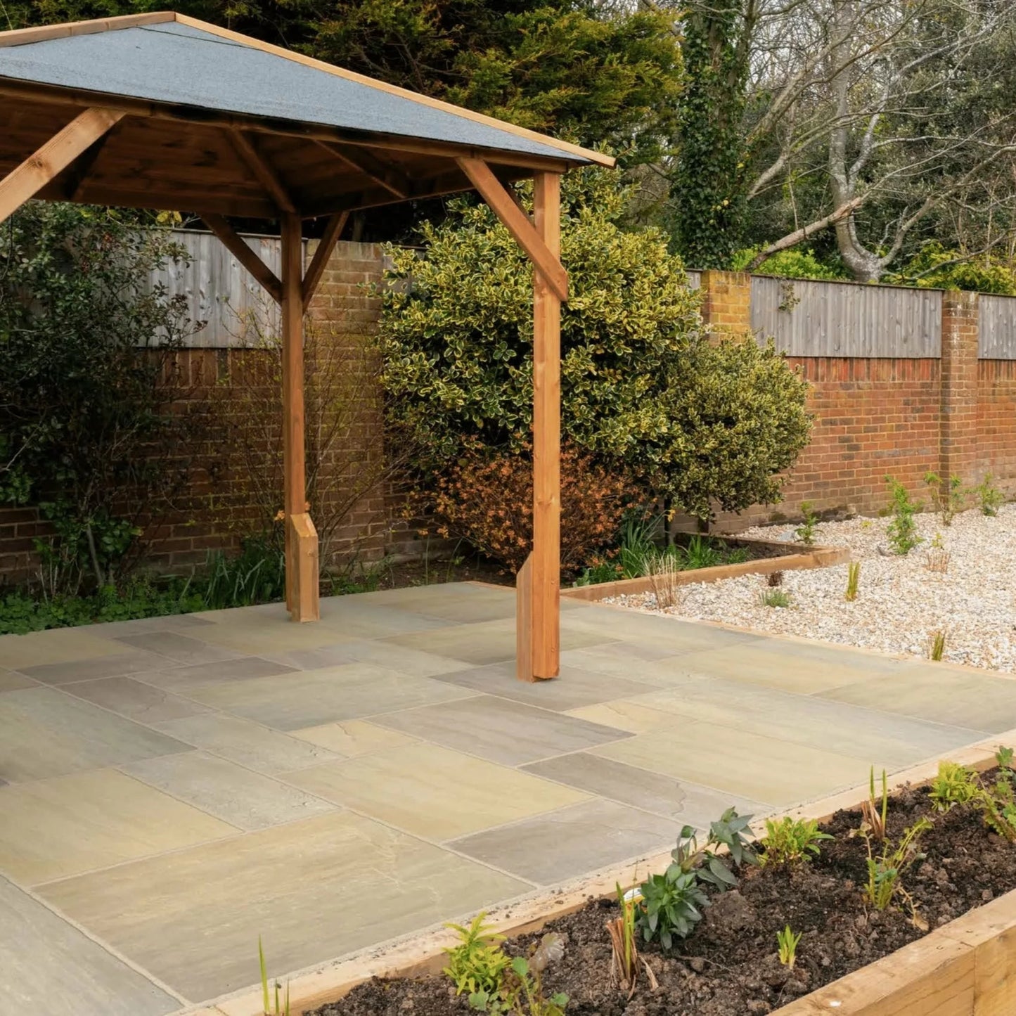 A small wooden gazebo stands on a paved patio of Raj Green Sandstone Paving in a backyard garden, surrounded by plants and gravel, bordered by a brick wall and wooden fencing. Trees and shrubs are visible in the background.