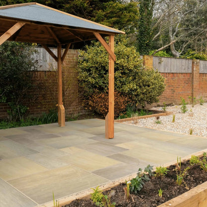 A small wooden gazebo stands on a paved patio of Raj Green Sandstone Paving in a backyard garden, surrounded by plants and gravel, bordered by a brick wall and wooden fencing. Trees and shrubs are visible in the background.