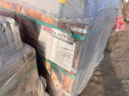A pallet wrapped in plastic contains sandstone slabs. A label reads "Kandla Grey Sandstone Patio Kit," alongside some handwritten numbers. The pallet, surrounded by paving tiles, sits on rough dirt ground outdoors.