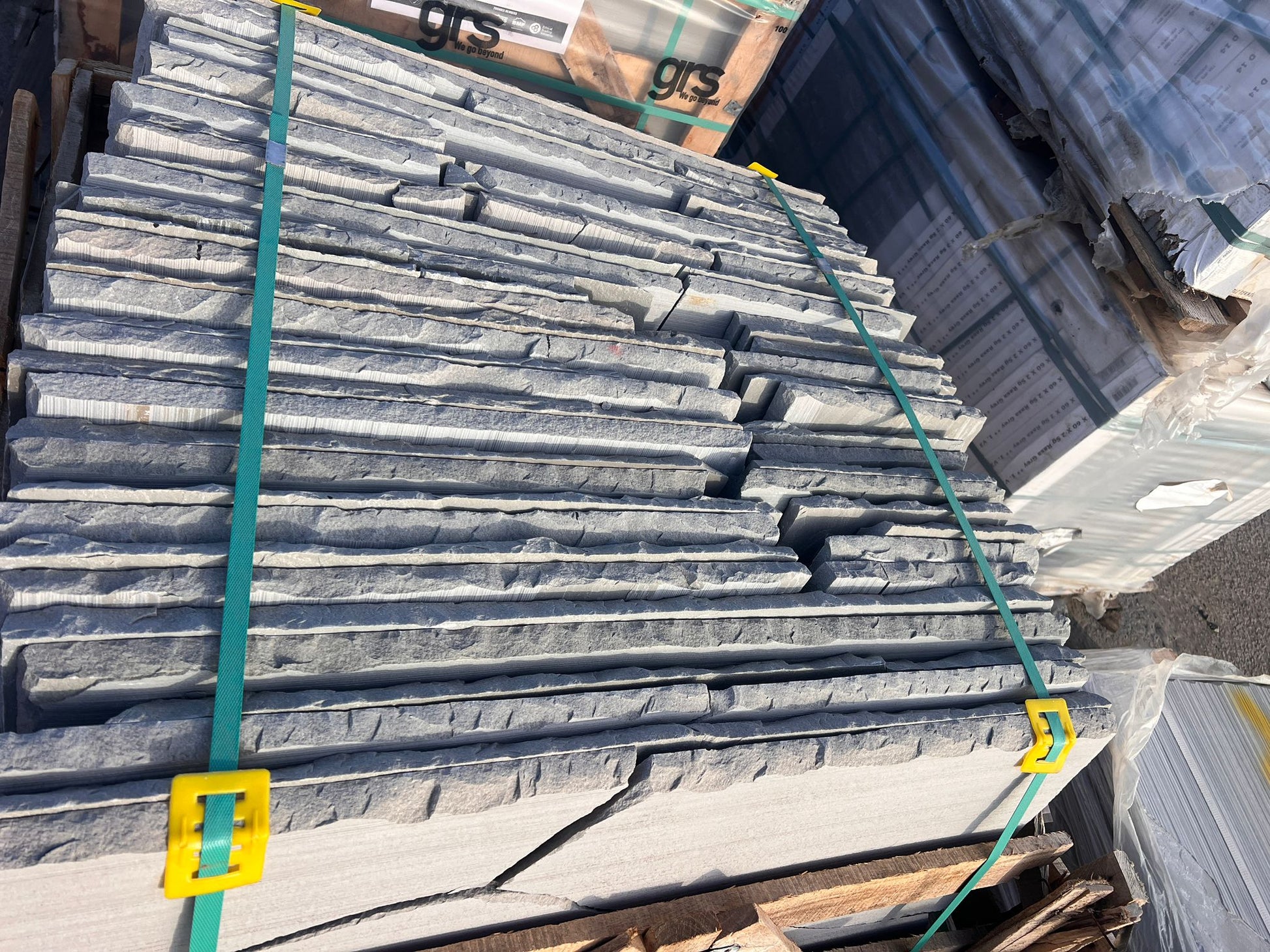 Stacks of thin, rough-edged Autumn Brown Sandstone Paving slabs bundled with green straps and yellow clasps, placed on a pallet outdoors among other building materials.