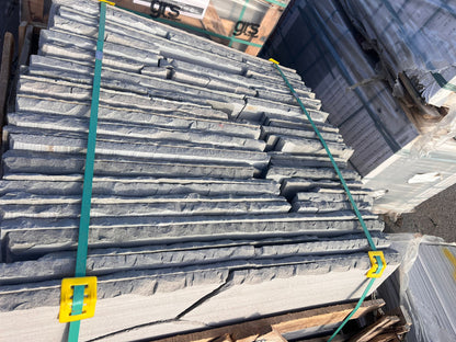 Stacks of thin, rough-edged Autumn Brown Sandstone Paving slabs bundled with green straps and yellow clasps, placed on a pallet outdoors among other building materials.