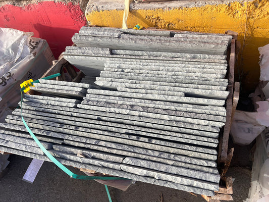 A stack of rectangular Autumn Brown Sandstone Paving slabs with textured surfaces is placed on a wooden pallet outdoors, against a painted red and yellow wall. Green straps are draped across the sandstone slabs.