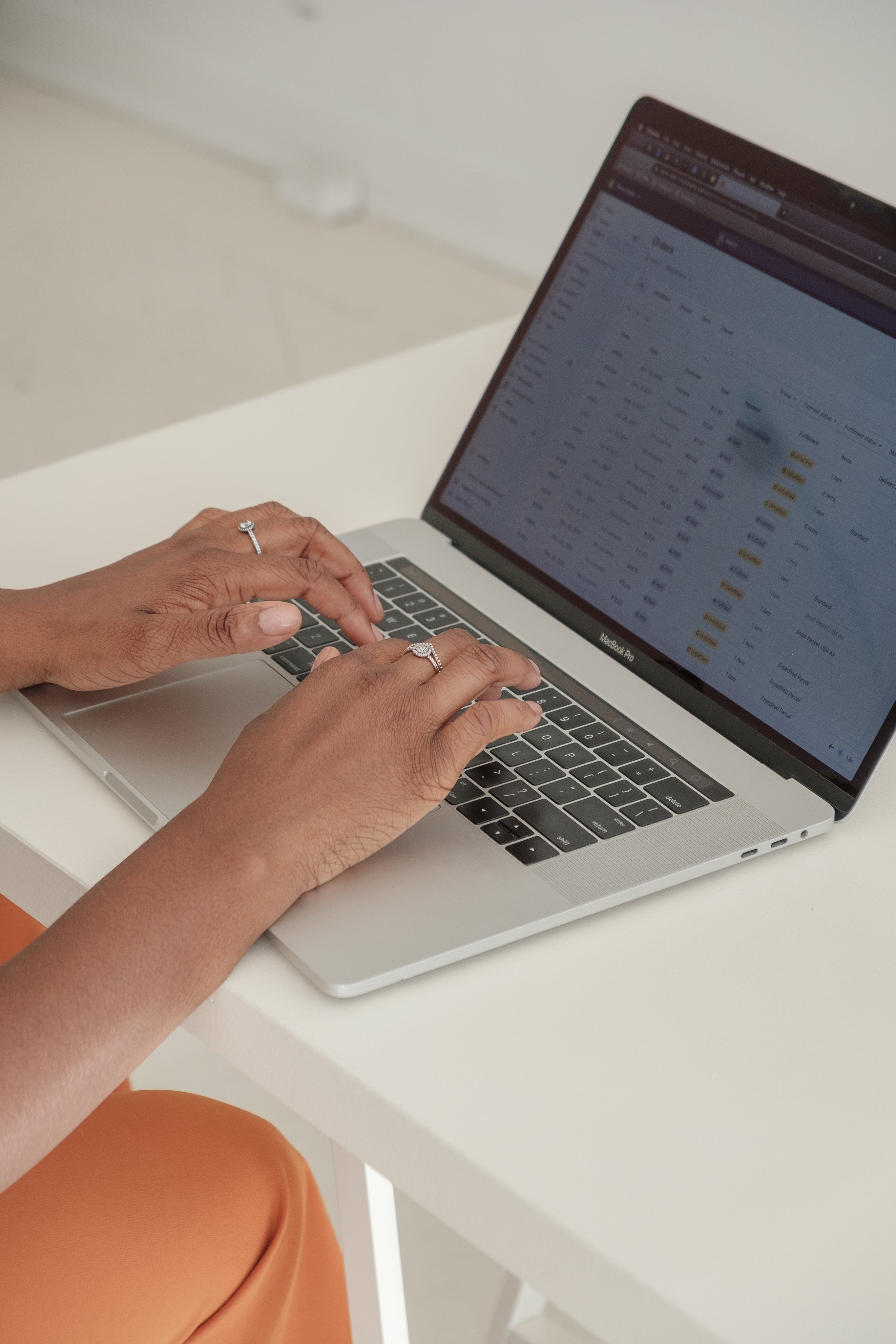 A person types on a laptop displaying a spreadsheet with rows and columns. The person's hands are visible, adorned with rings, and they are seated at a white desk.