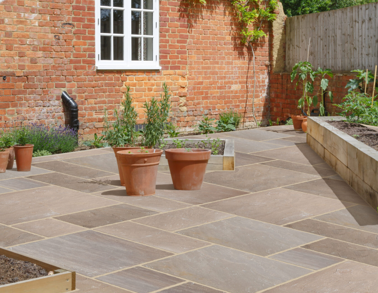 A paved garden patio with large beige tiles, terracotta pots with small plants, raised wooden beds, and a red brick house with a white-framed window in the background.