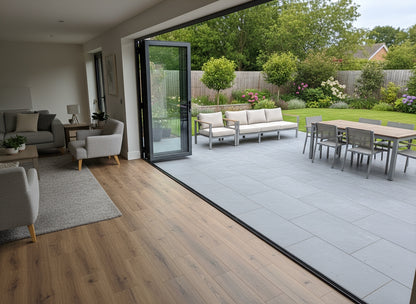 A modern living room with light gray furniture opens onto a patio featuring slip-resistant tiles and a dining table, overlooking a green garden with trees, shrubs, and flowering plants.