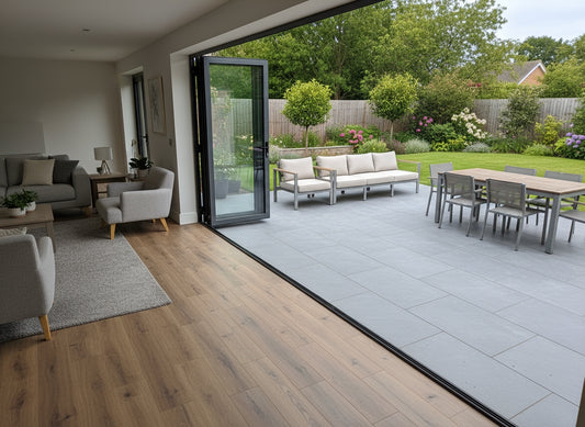A modern living room with light gray furniture opens onto a patio featuring slip-resistant tiles and a dining table, overlooking a green garden with trees, shrubs, and flowering plants.